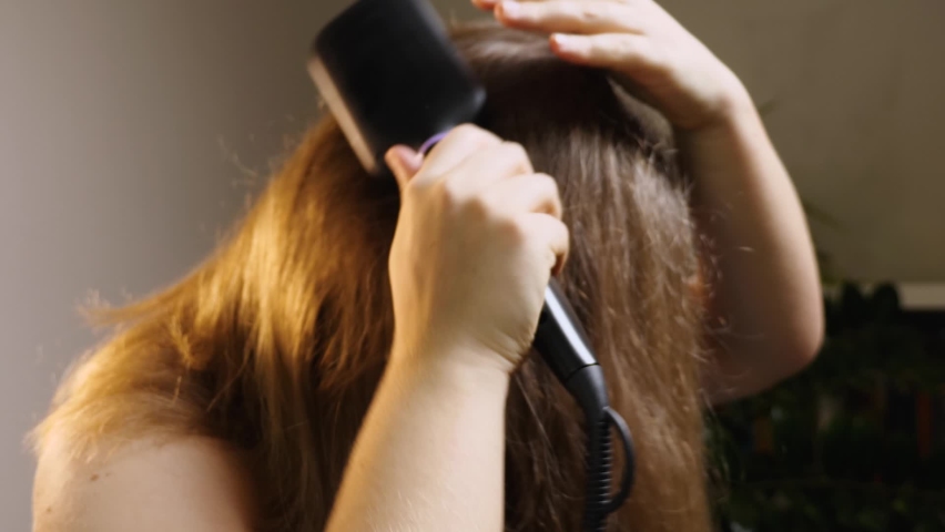 A girl standing in a room brushes her hair with a special electric straightening brush. The ceramic heated brush does not damage the hair. Close-up Royalty-Free Stock Footage #1088902191