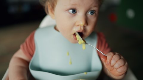 Closeup of young baby in white feeding high chair, kid is trying to eat himself, happy child with food stained face, little boy eats porridge with a spoon. High quality 4k footage