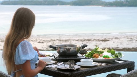 Woman cooking shabu shabu in hot pot on stove in Japanese restaurant with sea view. Various type of meat, seafood, vegetables on table for traditional asian dish sukiyaki