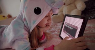The 7-year-old girl is playing in the children's room in unicorn costume and using digital computer tablet, enjoying cool video or photo content in social network, playing online games.