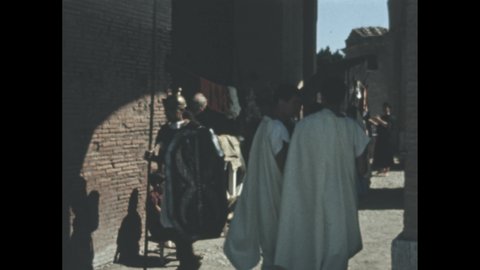 1960s: Man guides donkey through alley. Little boy sits in chair on side of alley. Roman style soldiers walk through alley. Sheep move around on hillside, shepherd stands near sheep.