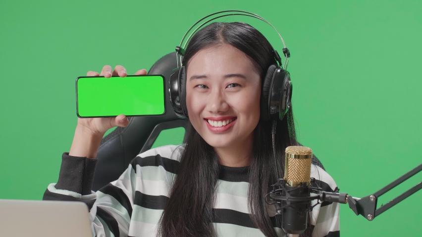 Close Up Of A Smiling Asian Woman With Headphone And Computer Showing Green Screen Mobile Phone While Sitting In Front Of Green Screen Background
