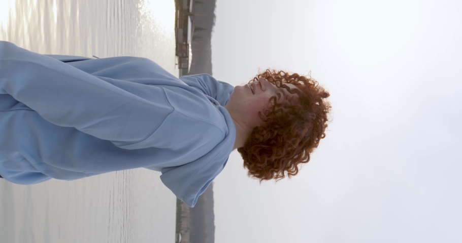 Red-haired charismatic plump girl with freckles waves her curly hair straightens her hand smiles looks into the camera at sunset against the backdrop of a lake in summer. vertical video | Shutterstock HD Video #1088916787