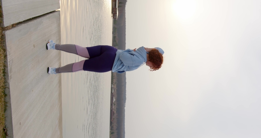 Plump girl curly red hair stands by the lake at sunset and does exercises by stretching her arms to her shoulders and lower back. floating departure of the camera from the steadicam. vertical video | Shutterstock HD Video #1088916815