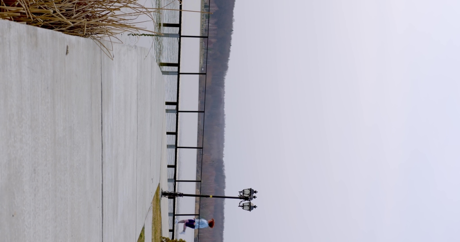 a curly-haired girl in sportswear runs along a wooden bridge on the lake opposite the forest, stops and does exercises, stretches her legs, in the foreground there is a fern. vertical view