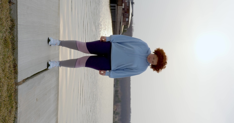 a beautiful curly girl with red hair in leggings and a blue bike stretches her neck, scrolling her head to the sides, doing an exercise at sunset on the lake. steadicam. vertical view