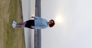 a curly-haired girl with red hair in leggings and a blue bike jumps and simultaneously raises her hands and claps her hands above her head on the lawn against the backdrop of the lake. vertical view