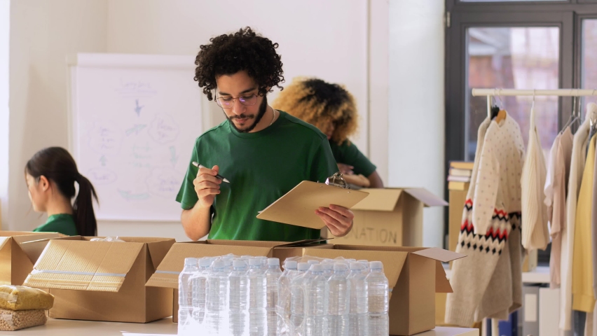 Charity, donation and volunteering concept - male volunteers with clipboard packing food and drinks in boxes over international group of people at distribution or refugee assistance center | Shutterstock HD Video #1088917345