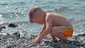 Cute baby boy playing on the seashore with rocks and pebbles. The kid is having a good time on a family vacation. Slow motion video