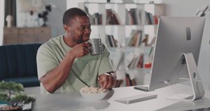 African american positive man staying home in casual apartment. Candid male watching fun internet videos, eating popcorn and drinking morning coffee.