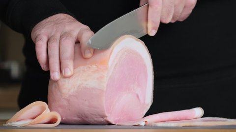 Prosciutto cotto with lard. Slicing traditional ham into pieces on cutting board with a knife. Thin slices of meat. Concept of delicious food snacks and cooking. Man with knife is cutting slim slices 