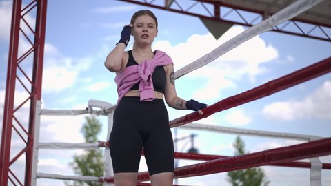 Tattooed female boxer entering boxing ring outdoors leaning on ropes looking away. Confident fit young Caucasian woman training on spring summer day. Healthy lifestyle and martial arts concept