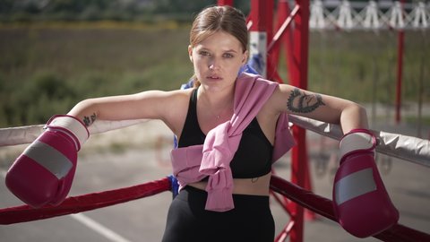 Portrait of confident female boxer posing in sunlight on boxing ring outdoors. Young motivated Caucasian tattooed woman sitting in sunshine looking at camera. Martial arts and sport concept