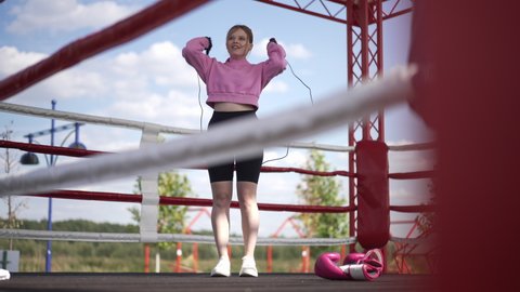 Wide shot motivated Caucasian sportswoman jumping rope in slow motion on boxing ring. Front view portrait of confident inspired woman working out training in sunshine outdoors. Sport concept