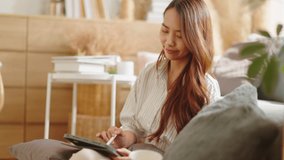 Smiling Asian freelance business women casual wear sitting on sofa using tablet writing note,plan,message and working call video conference in living room.Happy young girl relax at home in summer