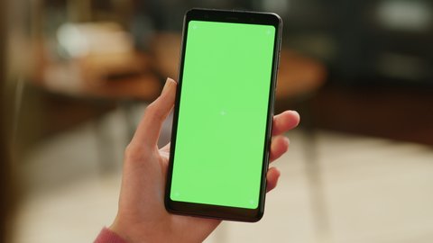 Feminine Hand Holding a Smartphone with Green Screen Mock Up Display. Female is Resting at Home, Watching Videos and Reading Social Media Posts on Mobile Device. Close Up Over the Shoulder Footage.