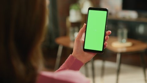 Feminine Hand Holding a Smartphone with Green Screen Mock Up Display. Female in Casual Clothes Resting at Home and Watching Videos on Mobile Device. Close Up Over the Shoulder Footage.