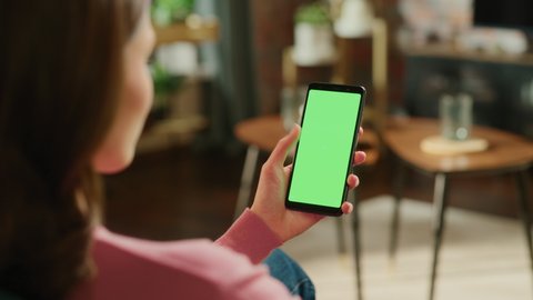 Feminine Hand Scrolling Feed on Smartphone with Green Screen Mock Up Display. Female in Casual Clothes Resting at Home and Checking Social Media on Mobile Device. Close Up Over the Shoulder Footage.