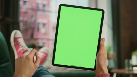 Feminine Hand Swiping Feed on Tablet with Green Screen Mock Up Display. Female is Relaxing on Sofa at Home, Watching Videos and Reading Social Media Posts on Mobile Device. Close Up POV Footage.