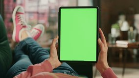 Feminine Hand Vertically Holding a Tablet with Green Screen Display. Female is Relaxing on a Couch at Home, Watching Videos and Reading Social Media Posts on Mobile Device. Close Up POV Footage.