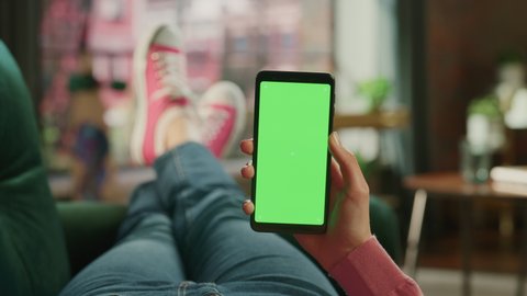 Feminine Hand Scrolling Feed on Smartphone with Green Screen Mock Up Display. Female is Relaxing on Sofa at Home, Watching Videos and Reading Social Media Posts on Mobile Device. Close Up POV Footage.