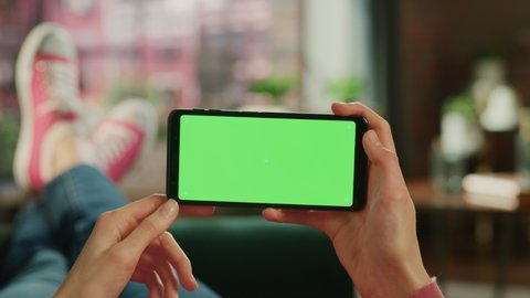 Woman Horizontally Holding a Smartphone with Green Screen Mock Up Display. Female is Relaxing on a Couch at Home, Watching Videos and Reading Social Media Posts on Mobile Device. Close Up POV Footage.