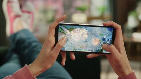 Female is Relaxing on a Couch at Home, Playing an Interactive PvP RPG Strategy Video Game on Her Smartphone. Gamer Lies on a Sofa in Living Room. Close Up POV Footage of Mobile Device Screen.