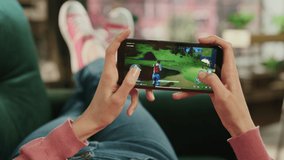 Female is Relaxing on a Couch at Home, Playing an Interactive PvP Shooter Video Game on Her Smartphone. Gamer Lies on a Sofa in Living Room. Close Up POV Footage of Mobile Device Screen.