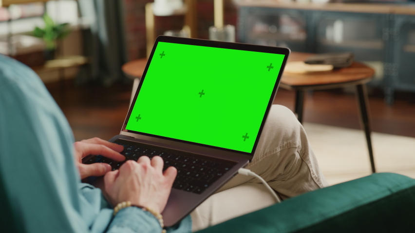 Young Adult Man Working from Home Loft Apartment on Laptop Computer with Green Screen Mock Up Display. Creative Male Browsing the Web, Writing on Social Media. Close Up Over the Shoulder Footage. Royalty-Free Stock Footage #1088937097