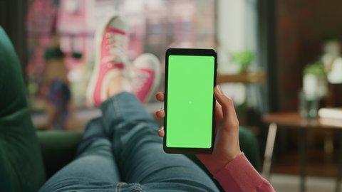 Feminine Hand Holding a Smartphone with Green Screen Mock Up Display. Female is Relaxing on a Couch at Home, Watching Videos and Reading Social Media Posts on Mobile Device. Close Up POV Footage.