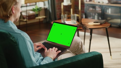 Young Adult Man Working from Home Loft Apartment on Laptop Computer with Green Screen Mock Up Display. Creative Male Browsing the Web, Writing on Social Media. Close Up Over the Shoulder Footage.