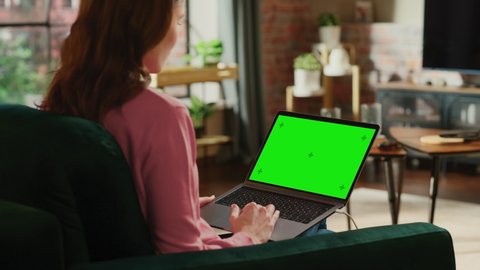 Young Adult Woman Working from Home Loft Apartment on Laptop Computer with Green Screen Mock Up Display. Creative Female Using Trackpad and Browsing Internet. Close Up Over the Shoulder Footage.