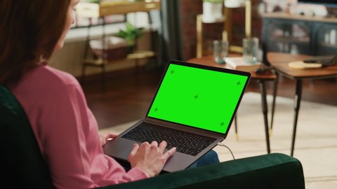 Young Adult Woman Working from Home Loft Apartment on Laptop Computer with Green Screen Mock Up Display. Creative Female Using Trackpad and Browsing Internet. Close Up Over the Shoulder Footage.