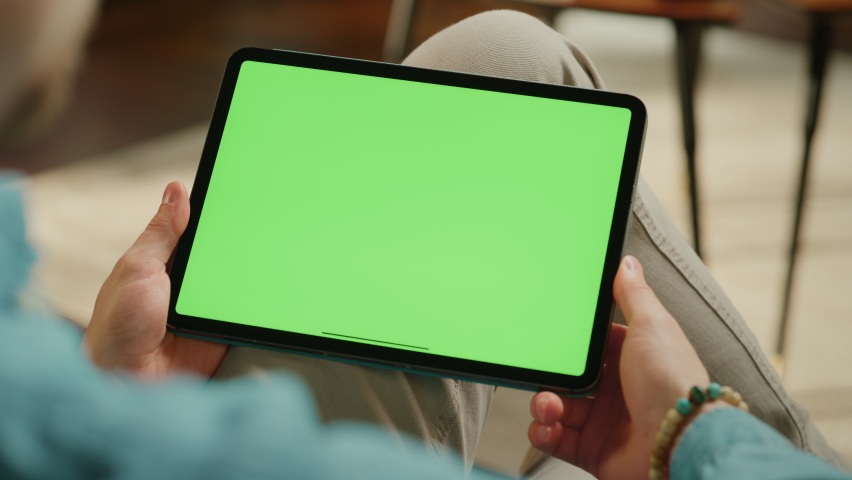 Young Man Scrolling and Tapping on Content on Tablet Computer with Green Screen Mock Up Display. Male Relaxing at Home, Reading Social Media Posts on Mobile Device. Close Up Over the Shoulder Footage.