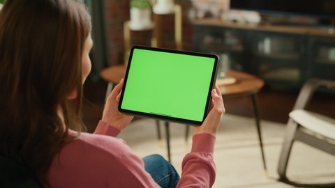 Young Woman Holding Tablet Computer with Green Screen Mock Up Display. Female Resting at Home, Watching Videos and Reading Social Media Posts on Mobile Device. Close Up Over the Shoulder Footage.