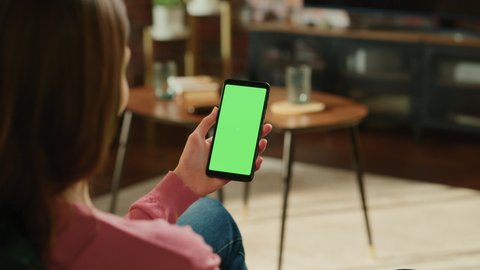 Feminine Hand Holding a Smartphone with Green Screen Mock Up Display. Female is Relaxing at Home, Watching Videos and Reading Social Media Posts on Mobile Device. Close Up Over the Shoulder Footage.