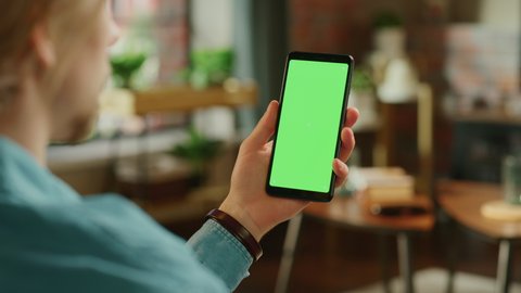 Man Holding a Smartphone with Green Screen Mock Up Display. Male is Resting at Home, Watching Videos and Reading Social Media Posts on Mobile Device. Close Up Over the Shoulder Footage.