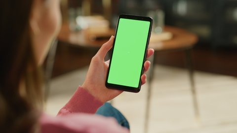 Feminine Hand Tapping on Display and Scrolling Feed on Smartphone with Green Screen Mock Up Display. Female Resting at Home, Checking Social Media on Mobile Device. Close Up Over the Shoulder Footage.