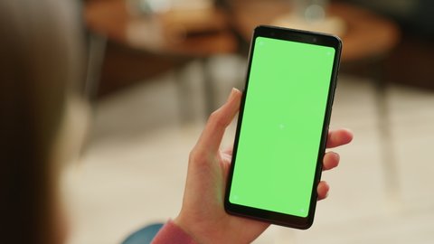 Feminine Hand Scrolling Feed on Smartphone with Green Screen Mock Up Display. Female is Resting at Home and Checking Social Media on Mobile Device. Close Up Over the Shoulder Footage.