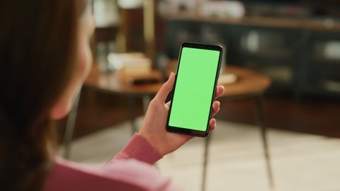 Feminine Hand Holding a Smartphone with Green Screen Mock Up Display. Female is Resting at Home, Watching Videos and Reading Social Media Posts on Mobile Device. Close Up Over the Shoulder Footage.