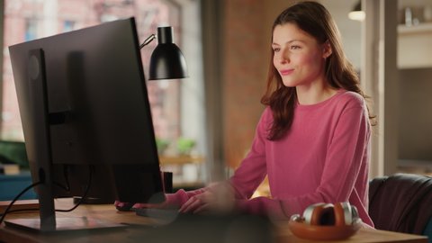Young Beautiful Adult Woman in Pink Jumper Working from Home on Desktop Computer. Creative Female Checking and Writing Emails. Loft Apartment with Urban City View from Big Window.