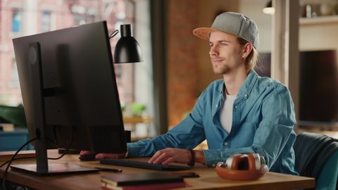 Young Handsome Adult Man Wearing a Cap, Working from Loft Apartment on Desktop Computer. Talented Male Checking and Writing Emails from Home or Creative Office. Urban City View from Big Window.