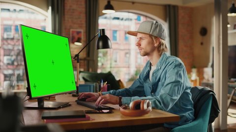 Young Handsome Adult Man Wearing a Cap, Working from Home on Desktop Computer with Green Screen Mock Up Display. Creative Male Checking and Writing Emails. Urban City View from Big Window.