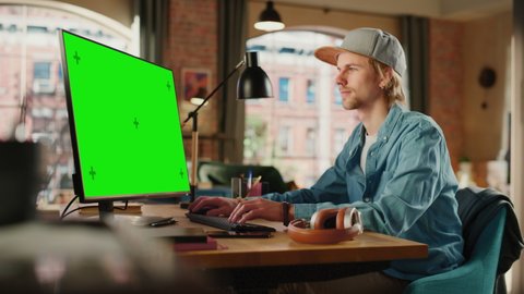 Young Handsome Man Working from Home on Desktop Computer with Green Screen Mock Up Display. Creative Male Checking Social Media, Browsing Internet. Urban City View from Big Window.