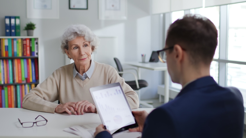 Senior woman meeting financial advisor and looking at charts on digital tablet. Back view of banker explain investment strategy to aged female client Royalty-Free Stock Footage #1088941043