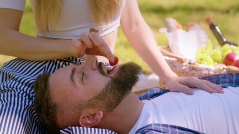 Love games at picnic. Relaxed bearded man lying on his lady knees, unrecognizable woman feeding her husband with fresh strawberry, resting in summer park together, slow motion