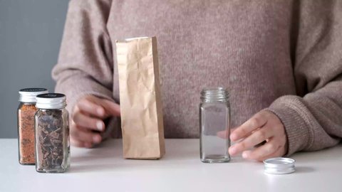 Unrecognizable woman refilling spice jar with dry cayenne pepper from a paper bag buyed at package free grocery store.