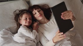 Beautiful young mom and her cute girl in white pajamas lying on the bed at home , laughing and looking in digital tablet. Mother and her daughter lying in bed, having fun with digital tablet. - Powered by Shutterstock - Get 15% off with code: PIKWIZARD15
