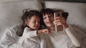 Mother and daughter lyuing on a coach in the bedroom at home and using mobile phone having a video call. Happy woman and her little daughter using mobile phone while relaxing together in bed at home.