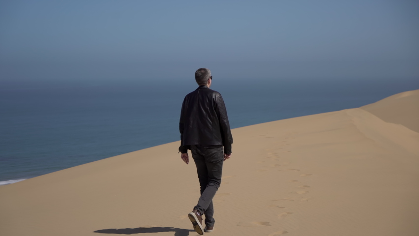A young man walks along sandy dunes on the seashore. Sandwich Harbour in Namibia. Summer vacations, wild desert nature in Africa. Coast of the Atlantic Ocean, sea water. Raise hands up, feel freedom. Royalty-Free Stock Footage #1088944087